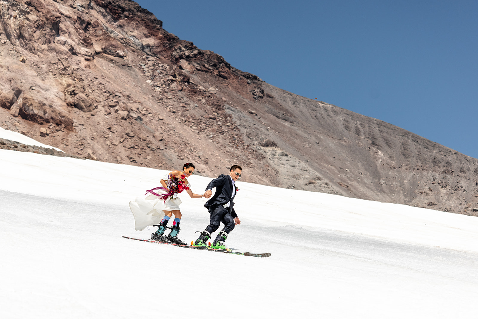 Skiing elopement, a couple holds hands while skiing down a slope in wedding clothes
